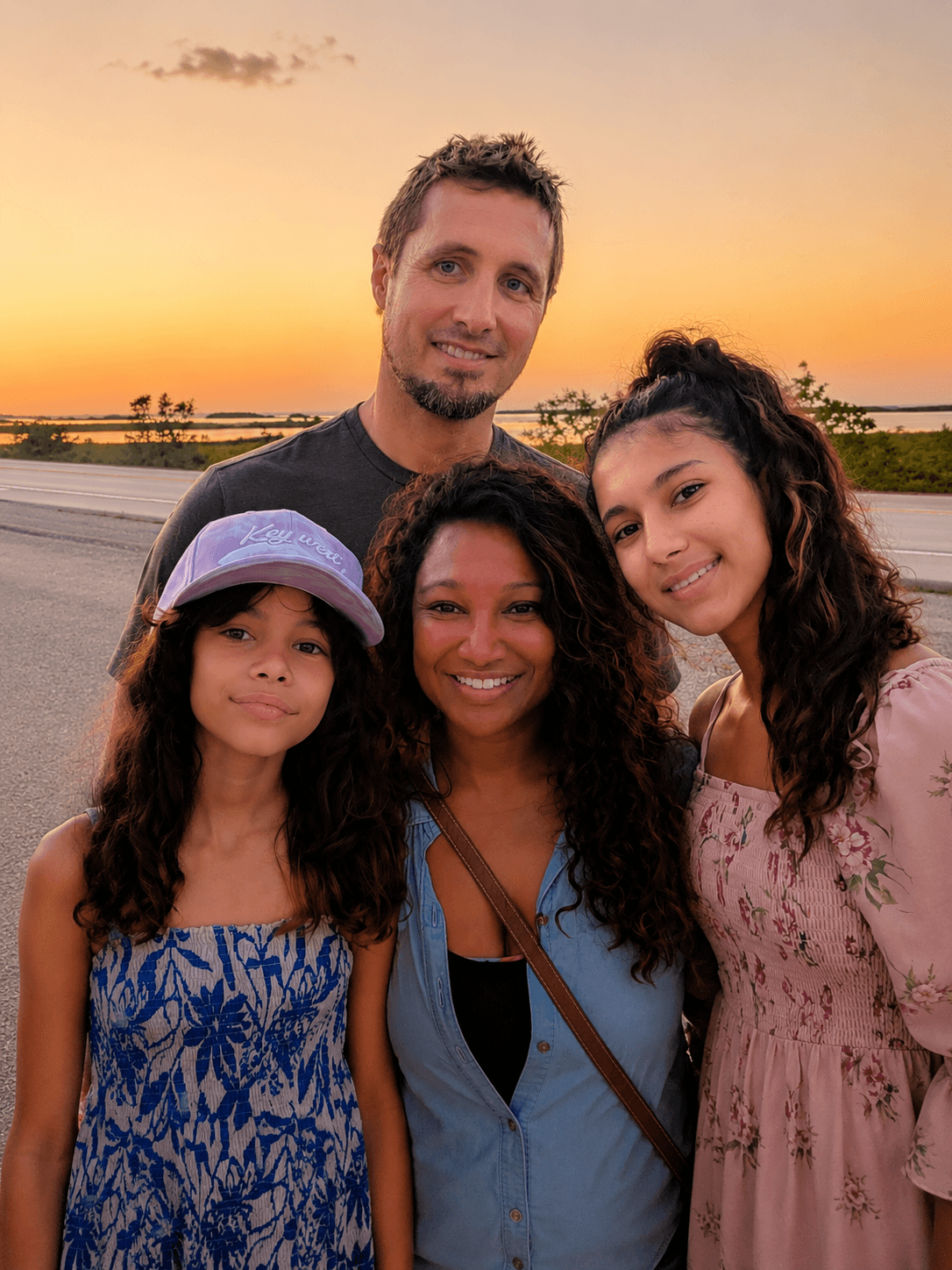 Smiling family of four posing outdoors against a vibrant orange sunset sky near the coast.